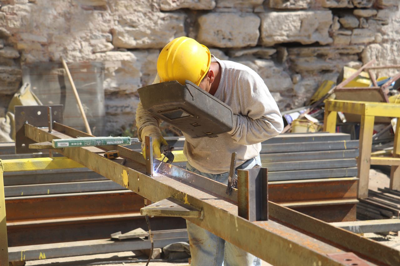 Welder wearing safety gear works with metal beams outdoors on a construction site.